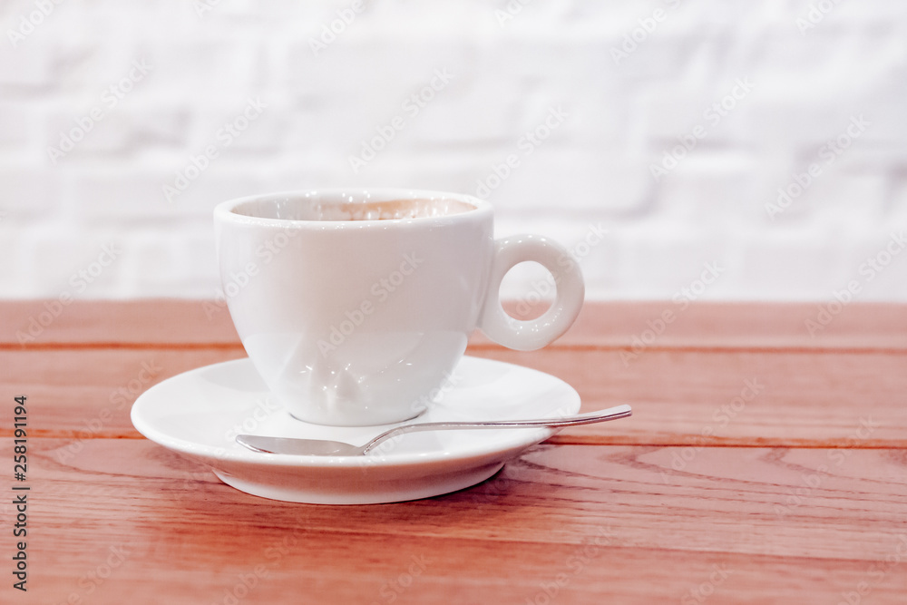 A white coffee cup with freshly brewed coffee and a spoon on a saucer stands on a dark wooden table near a white wall with a light shade on the table top in a cafe. Close up. A white coffee cup with freshly brewed coffee and a spoon on a saucer stands on a dark wooden table near a white wall with a light shade on the table top in a cafe. Close up.