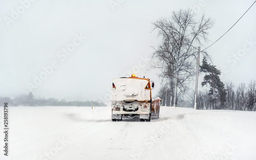 Orange plow truck on snow covered road, gray sky and trees in background - winter road maintenance