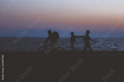 creative soft focus human wallpaper pattern of fuzzy walking people silhouette on dark twilight waterfront along Pacific ocean and unfocused mountain horizon background in evening sunset time