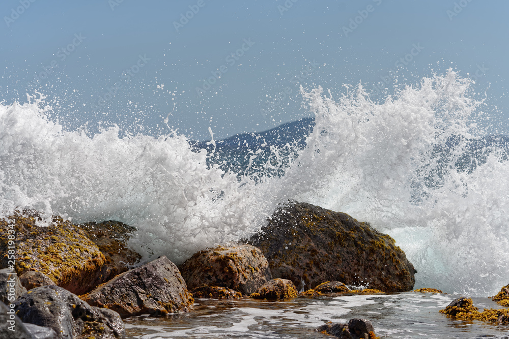 Obraz premium A wave breaks on big stones in the water, thousands of drops of water sparkle in the sunlight, island silhouette in the background, snapshot - Location: Caribbean, island Guadeloupe