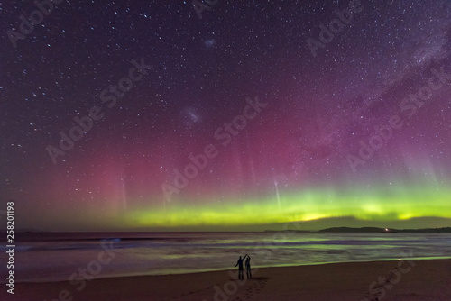 Two people in silhouette standing in awe of an incredible display of the Aurora Australis or Southern Lights, with bioluminescence turning the breaking waves bright blue.