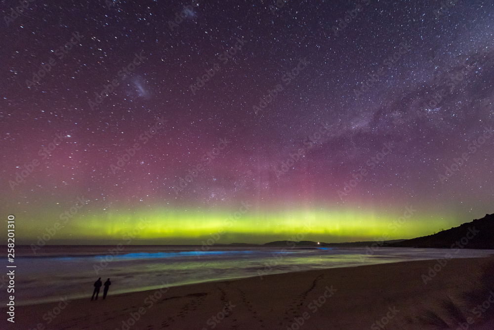 custom made wallpaper toronto digitalTwo people in silhouette standing in awe of an incredible display of the Aurora Australis or Southern Lights, with bioluminescence turning the breaking waves bright blue.