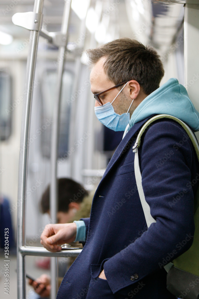 Fototapeta premium Exhausted man in eyeglasses feeling sick, wearing protective mask against transmissible infectious diseases and as protection against the flu in public transport/subway, people on background.