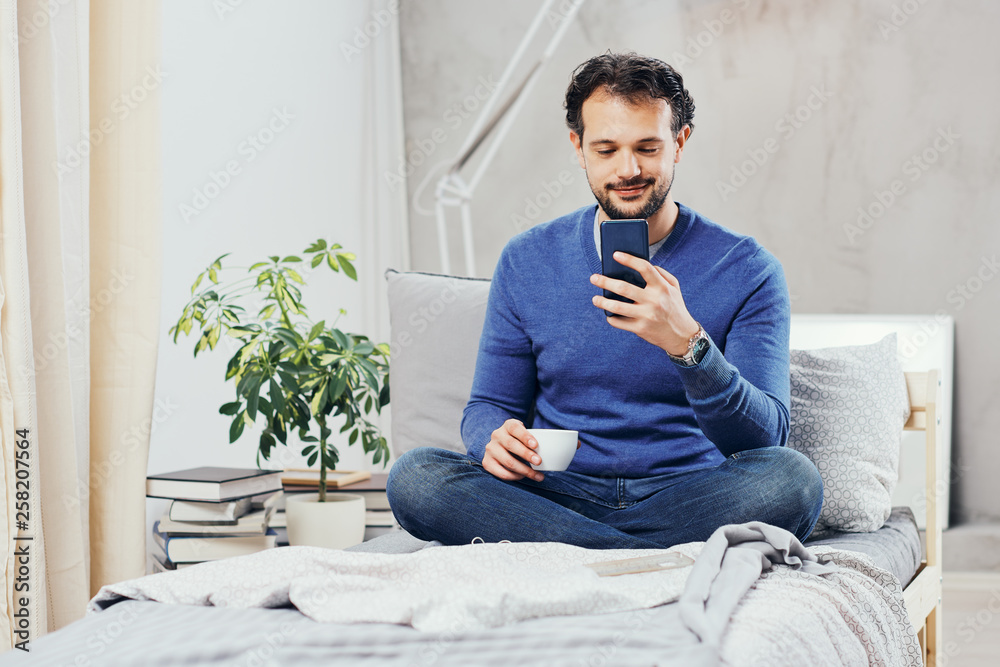 Attractive smiling arab man dressed casual sitting on bed in bedroom, drinking coffee and using smart phone for texting. Attractive smiling arab man dressed casual sitting on bed in bedroom, drinking coffee and using smart phone for texting.