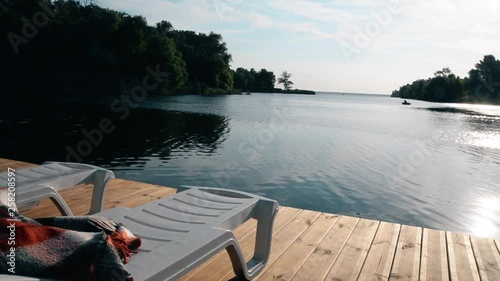 Long shot of lounge zone. Two loungers on a wooden pier on the broad river. Clear blue sky with bright sun and highlights.