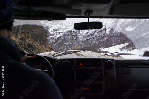 Fototapeta Naklejka Na Ścianę i Meble -  Inside view from the interior of a vehicle to the front window, with a winter route on the background with snow on the shoulder. The driver is in a blur and the interior is dark, with copy space