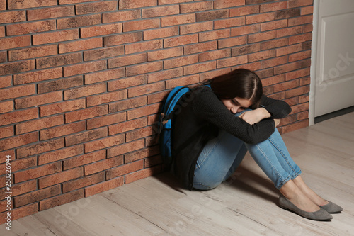 Upset teenage girl with backpack sitting on floor near wall. Space for text