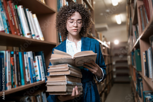 Young beautiful curly girl in glasses and a blue suit is standing in the library. Student Study