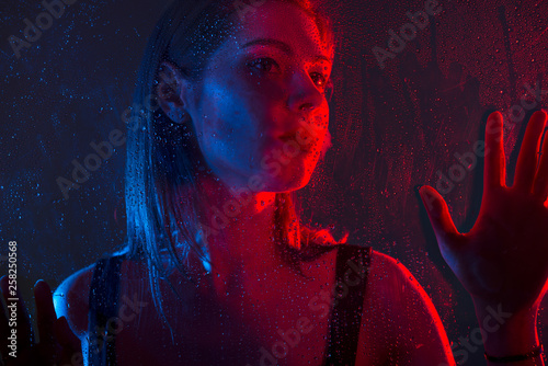 Glamorous brown-haired woman outside the window with drops of water in neon light close-up.
