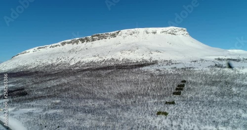Saana tunturi, C4k aerial, reverse, drone shot, of snowy finnish famous fell mountain, blue sky and arctic birches, on ground, on a sunny, winter day, in Enontekio, Lapland, Finland photo