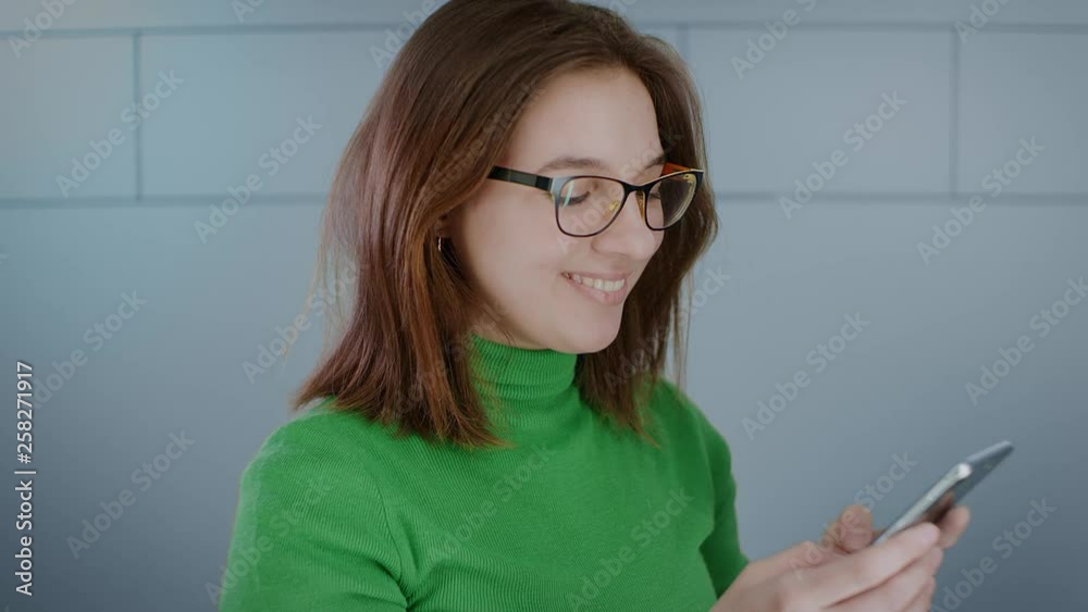 Attractive young girl with glasses typing a message on the phone, smiling, laughing looking at the phone screen. Brunette in a green jacket against a light wall. Close-up portrait. Slow motion. 4K Attractive young girl with glasses typing a message on the phone, smiling, laughing looking at the phone screen. Brunette in a green jacket against a light wall. Close-up portrait. Slow motion. 4K
