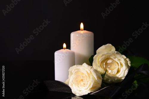Burning candles, mourning ribbon and flowers on black background
