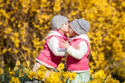 cute twin sisters, embrace and kissing on a background field with yellow flowers, happy cute and beautiful sisters having fun with yellow flowers in spring in park, cheerful holidays outdoors
