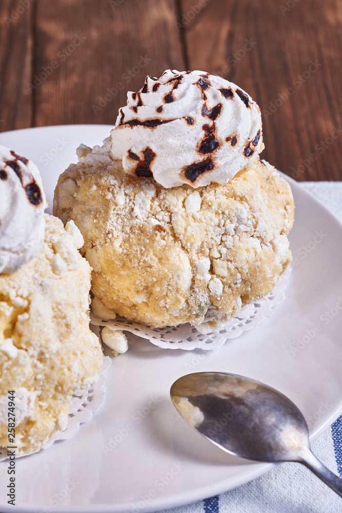 Two airy desserts with meringue in chocolate drops with prunes and butter cream in a white plate next to tea spoon and napkin on a wooden background. Close-up Two airy desserts with meringue in chocolate drops with prunes and butter cream in a white plate next to tea spoon and napkin on a wooden background. Close-up