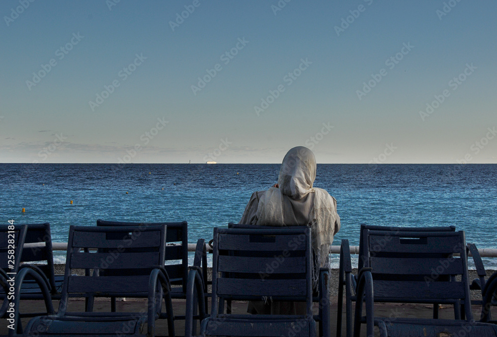 Woman with hijab sitting on a chair by the sea on Promenade des Anglaise in Nice, France. 