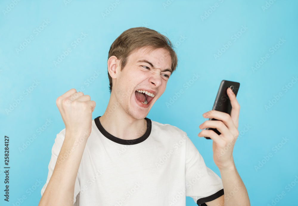 young funny male teenager in white t-shirt on blue background looks into smartphone as if won something young funny male teenager in white t-shirt on blue background looks into smartphone as if won something
