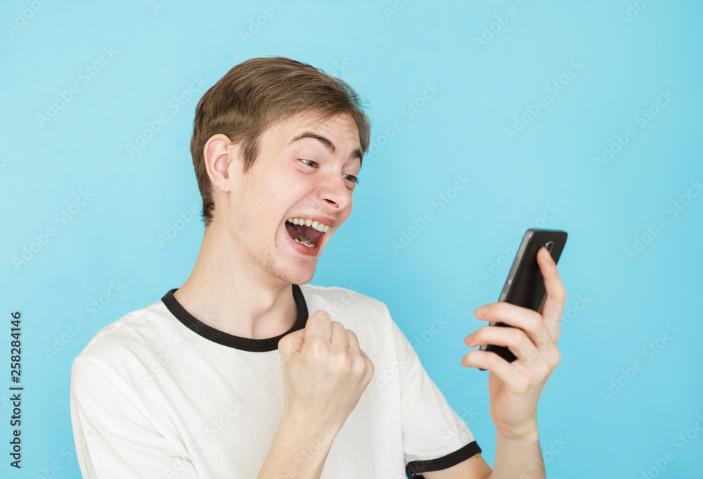 young funny male teenager in white t-shirt on blue background  looks into smartphone as if won something