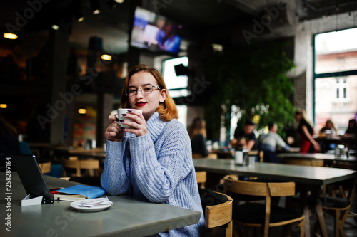 Cheerful young beautiful redhaired woman in glasses using her phone, touchpad and notebook while sitting at her working place on cafe with cup of coffee. © AS Photo Family