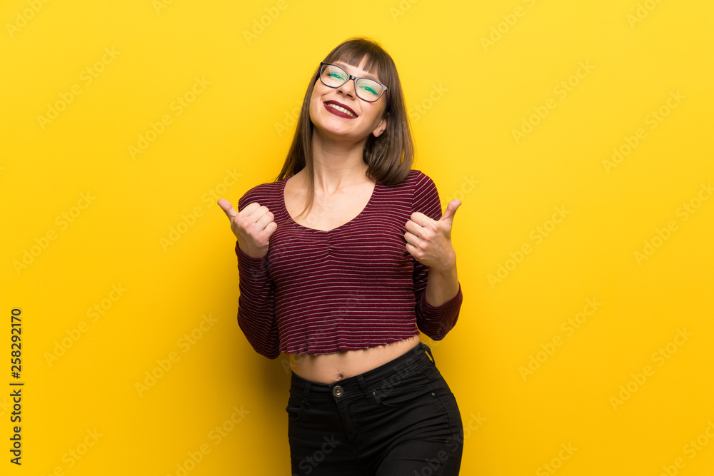 Woman with glasses over yellow wall giving a thumbs up gesture and smiling Woman with glasses over yellow wall giving a thumbs up gesture and smiling