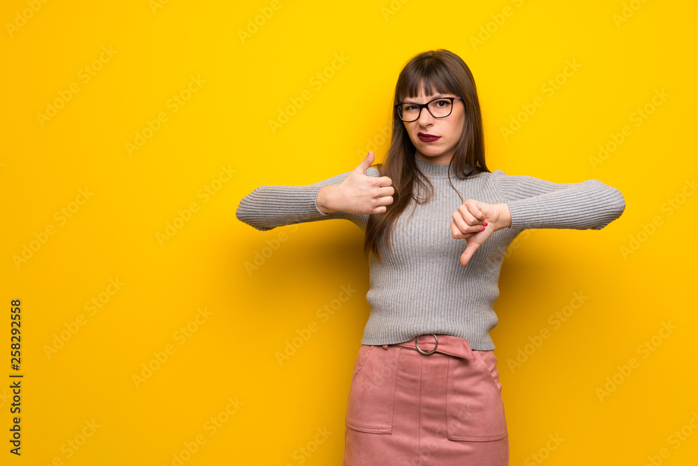Woman with glasses over yellow wall making good-bad sign. Undecided between yes or not Woman with glasses over yellow wall making good-bad sign. Undecided between yes or not