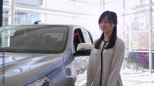 Auto business, portrait of smiling saleswoman pats car with pleasure and shows keys to new automobile for sale in sales center close-up photo