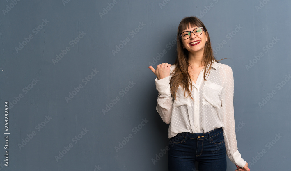 Woman with glasses over blue wall pointing to the side to present a product Woman with glasses over blue wall pointing to the side to present a product