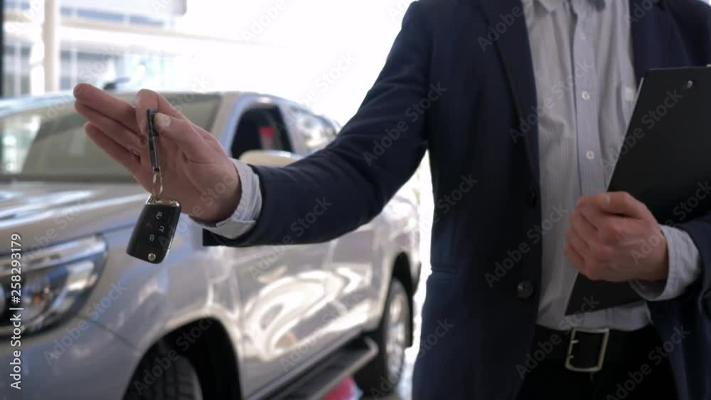 Auto business, young salesman holds in arms keys to new car for sale in sales center close-up Auto business, young salesman holds in arms keys to new car for sale in sales center close-up