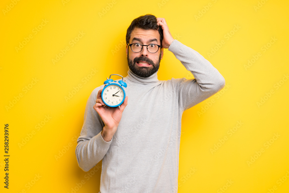 Man with beard and turtleneck restless because it has become late and holding vintage alarm clock - obrazy, fototapety, plakaty 