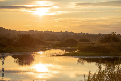 Beautiful sunset or sunrise golden landscape of river water, forest, sky and sun reflection on surface of water. Peaceful countryside and golden hours. Horizontal color photography.