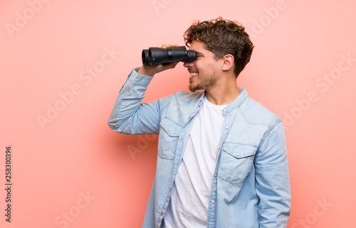 Blonde man over pink wall and looking in the distance with binoculars