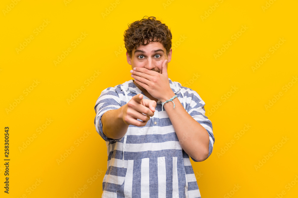 Blonde man over yellow wall pointing with finger at someone and laughing Blonde man over yellow wall pointing with finger at someone and laughing