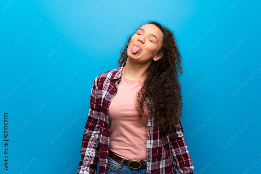 Fototapeta premium Teenager girl over blue wall showing tongue at the camera having funny look