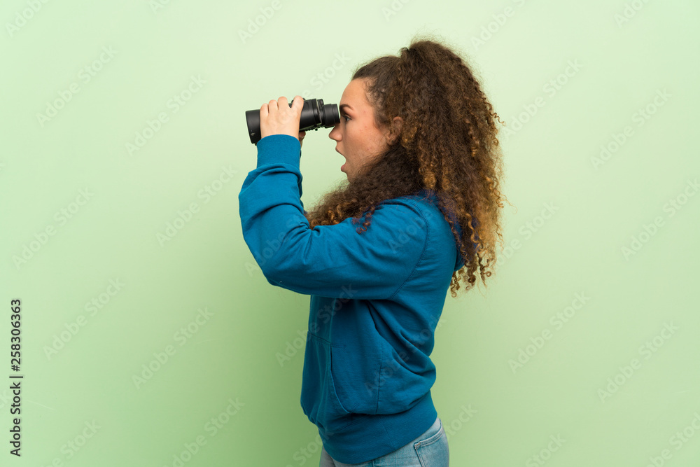 Obraz premium Teenager girl over green wall and looking in the distance with binoculars