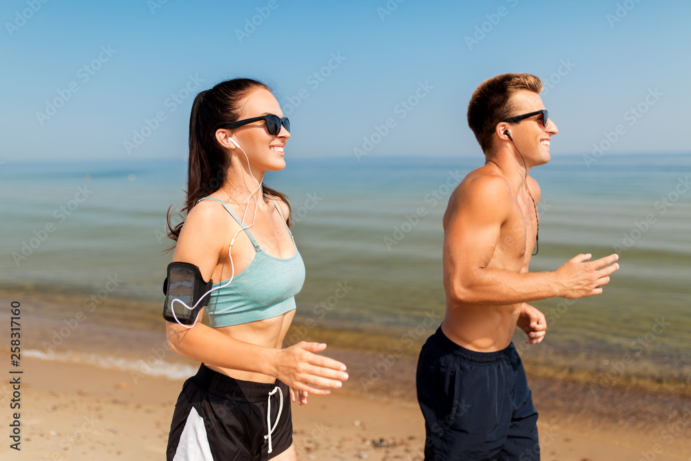 fitness, sport and technology concept - happy couple with earphones and arm bands running along summer beach
