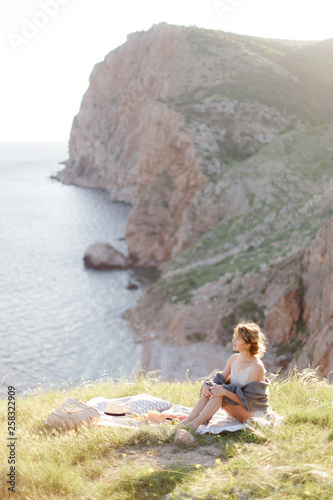 Young beautiful woman, wrapped in a blanket, sitting on the plaid and relaxing on nature. Picnic outdoors on the grass on mountain over sea view on sunny summer day.