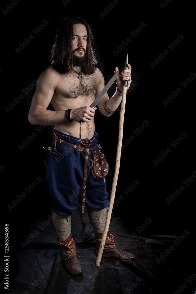 a strong bearded long-haired man with a tattoo on his shoulder is standing, holding in his hands a spear and knife of the Viking era. He is dressed with pants and shoes of 10-12 century