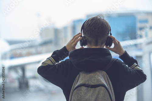 A boy in headphones with a backpack stands near the window at the airport and looks at the plane. Departure hall photo