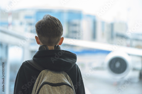 A boy in headphones with a backpack stands near the window at the airport and looks at the plane. Departure hall