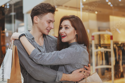 Cheerful loving young man hugging his beautiful girlfriend at the shopping mall. Young woman looking excited, while shopping at the mall with her boyfriend, copy space