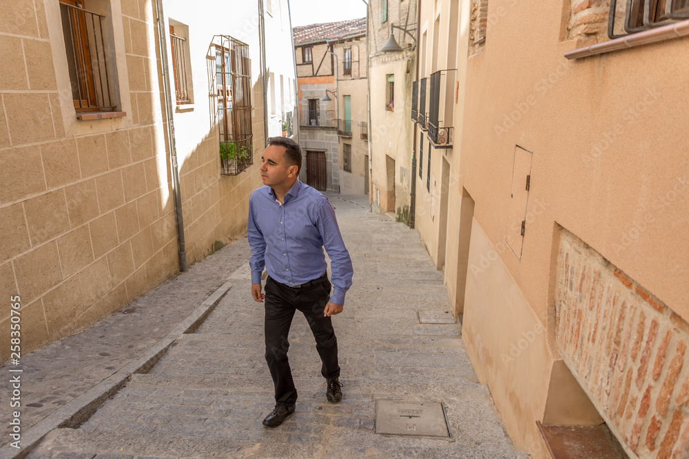 An elegant man in a blue shirt, walks through an old part of a city, on a sunny day