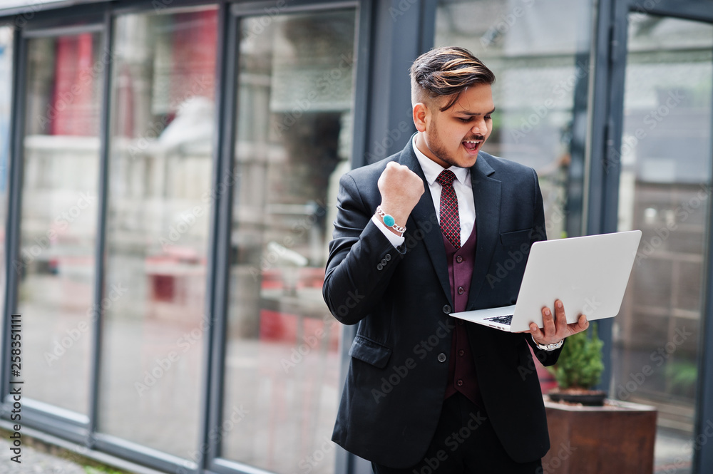 Fototapeta premium Stylish excited happy indian businessman in formal wear with laptop on hands standing against windows in business center. Gesture of the winner!