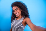 Closeup portrait of smiling young attractive African brazilian woman holding smartphone, taking selfie photo on the blue background.