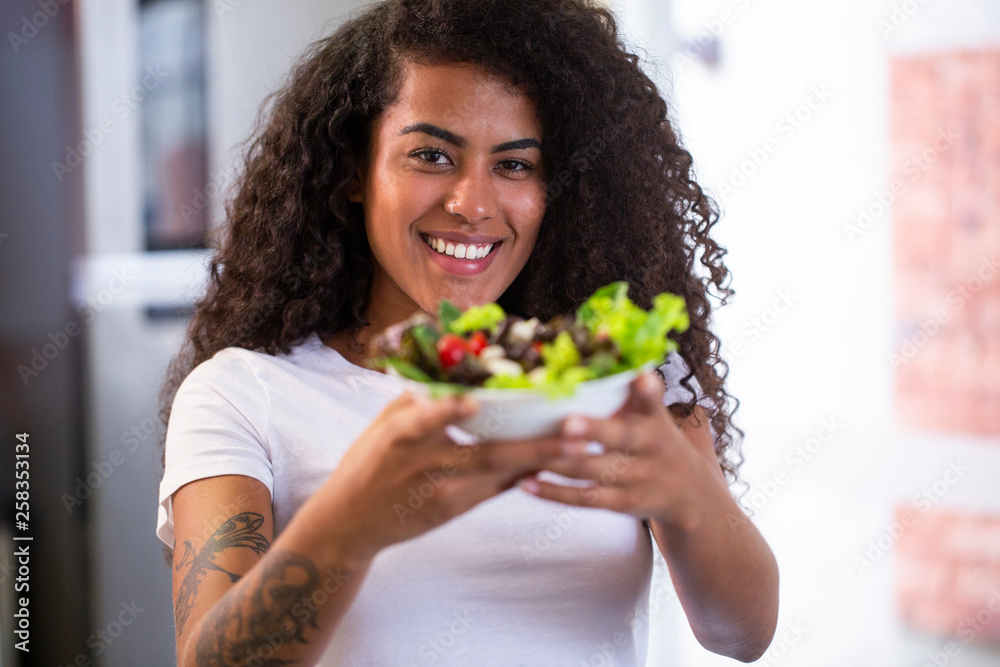cheerful young afro american woman eating vegetable salad in home kitchen - Imagem