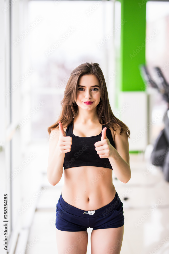 Portrait of happy sports lady standing and posing with thumbs up gesture in fitness club.