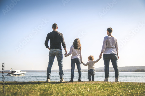 All family together holding hands near the sea. Four people standing on the seashore in a beautiful sunny day. Mother, father and two daughters looking at the horizon. Concept of friendship and union