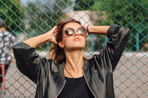 Fashion portrait of trendy young woman wearing sunglasses, and bomber jacket sitting next to rabitz in the city
