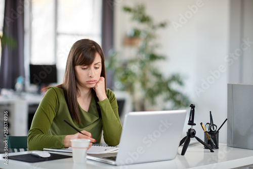 Charming Caucasian businesswoman sitting in moderna office and taking notes in notebook. In front of her laptop.
