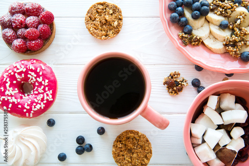 A cup of fresh coffee and tasty sweet snacks. Pink pastry and healthy cookies, pieces of banana and coconut. Coral color plates and cup on white wooden table. 