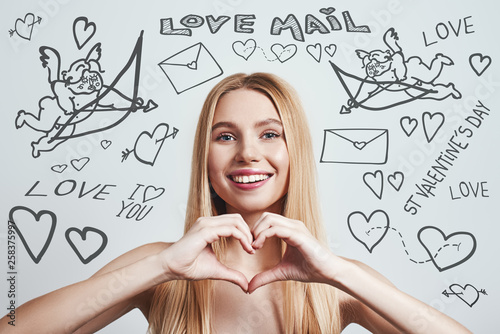 In love... Happy and smiling blonde woman making a romantic heart gesture with her hands while standing against grey background with doodles on the theme of St.Valentine's day. photo