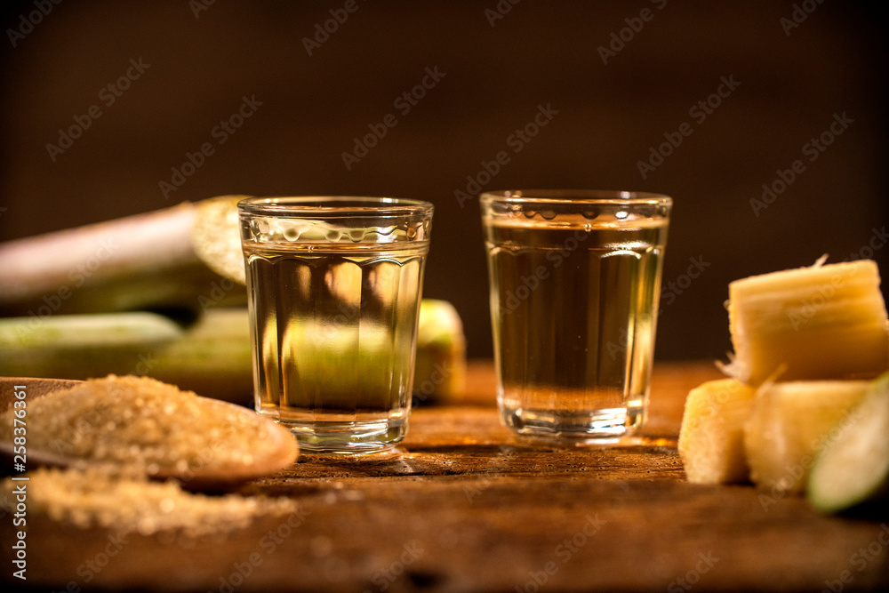 two shot glasses of Brazilian gold cachaca with sugar and sugarcane isolated on rustic wooden background